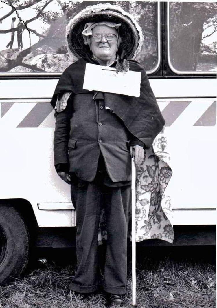 An elderly person in a suit, glasses, and a large hat stands with a white cane and patterned cloth draped over one shoulder, in front of a bus.