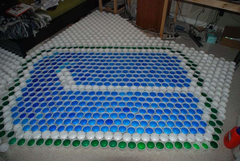 Hundreds of colored plastic yogurt cups arranged on the floor to form a large blue and white Gmail icon, outlined with green cups, in a cluttered room.
