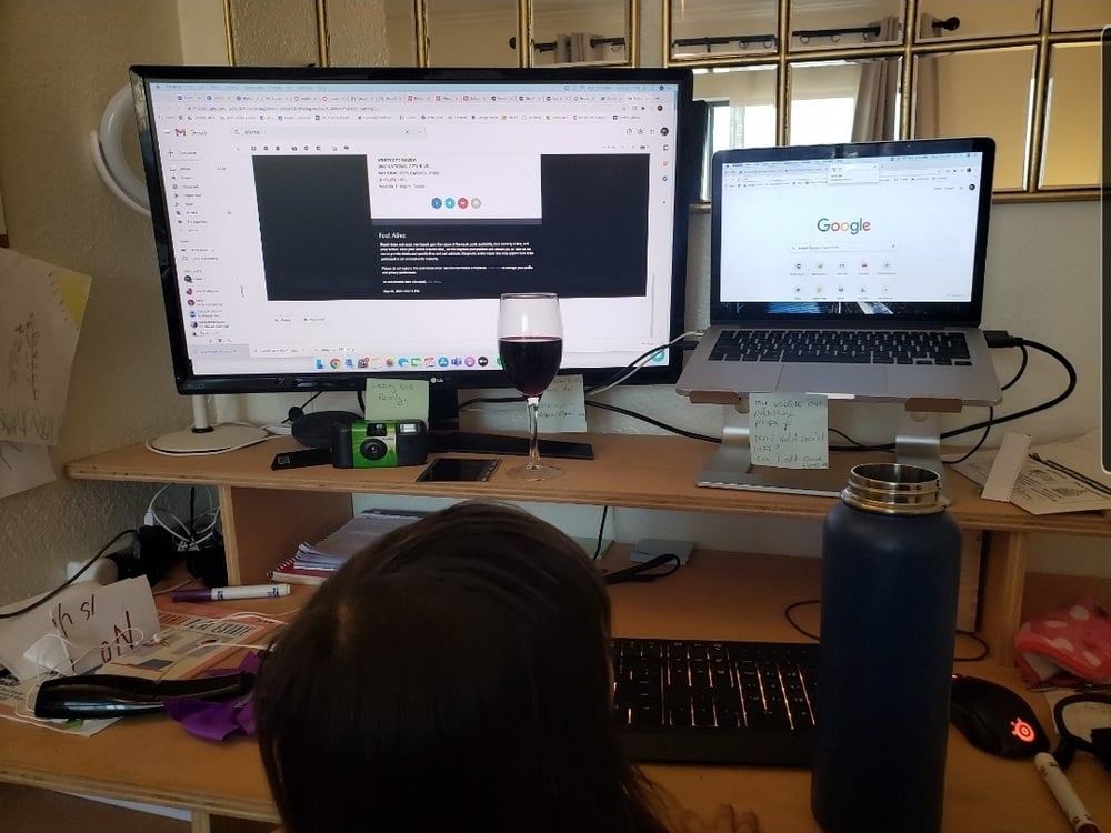 A child sits at a cluttered desk with dual monitors, a laptop on a stand, a glass of red liquid, a large blue bottle, and various papers and notes, suggesting a work-from-home setup.