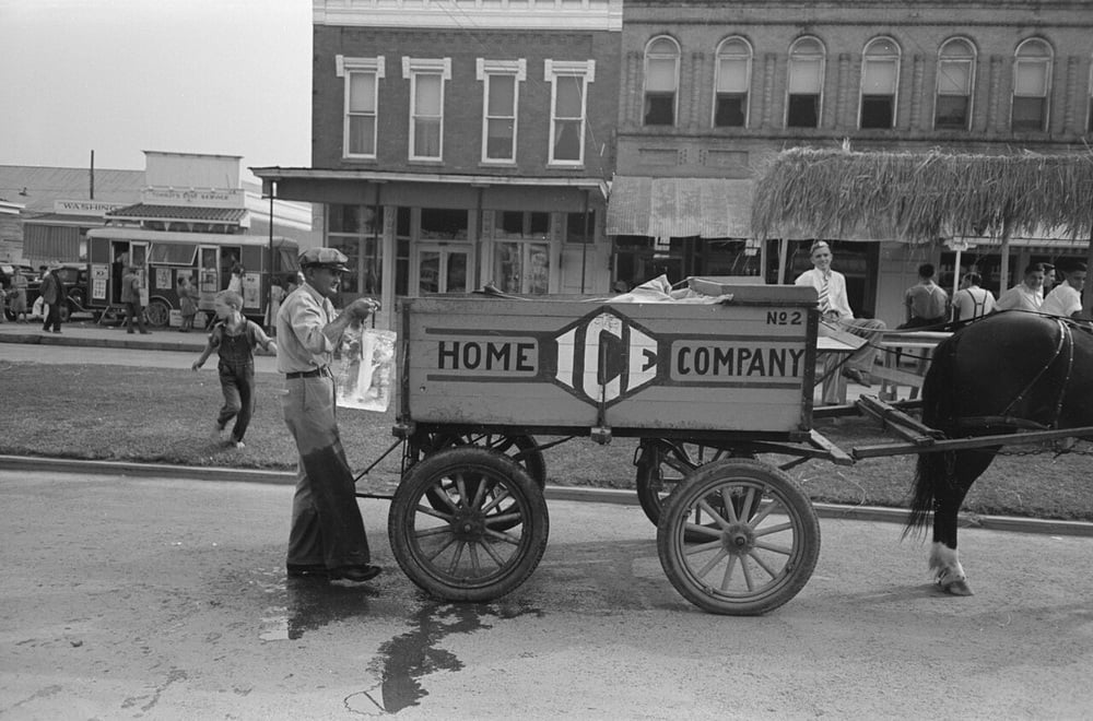 A man delivers ice from a wooden horse-drawn cart labeled “Home Ice Company” on a street with old-fashioned buildings. Children and other people are nearby.