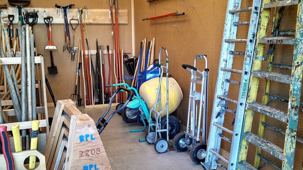 Shed interior with rakes, shovels, brooms, a wheelbarrow, ladders, hand trucks, and various yard tools neatly stored and organized along the walls.