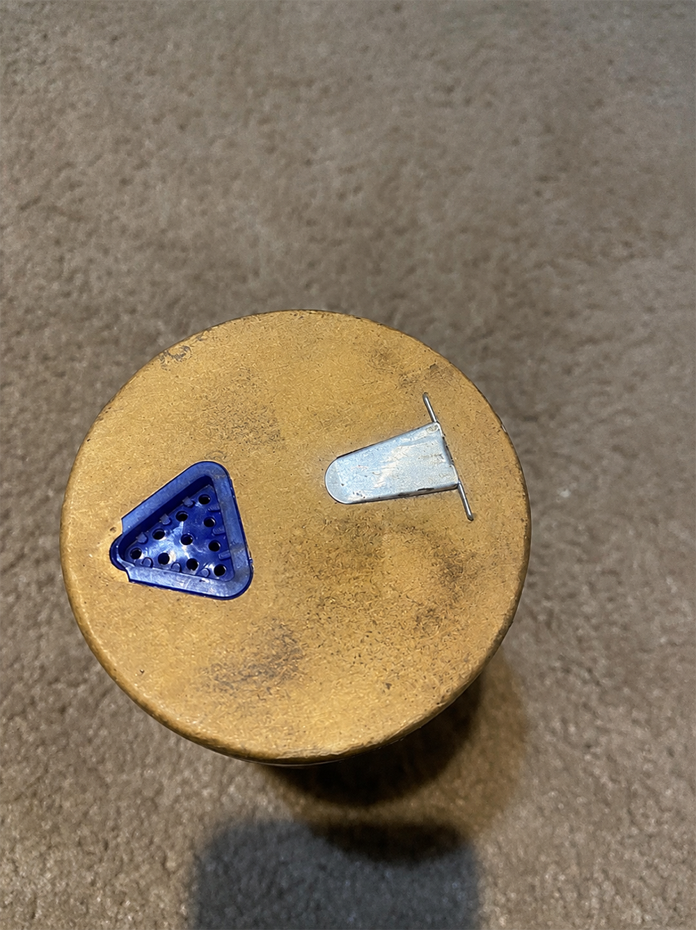 A brass cylinder with a blue triangular vent and a silver metal lever on top, placed on a brown carpeted floor.