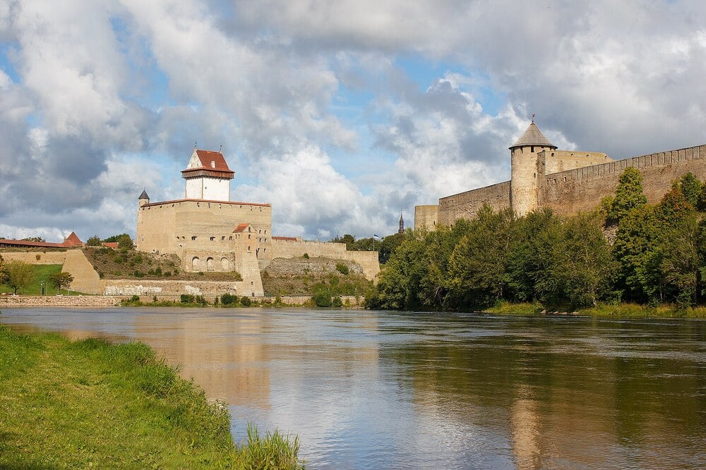 A historic stone fortress stands beside a calm river, with green trees and grass along the water’s edge. The sky above is partly cloudy, adding contrast to the scenic landscape.