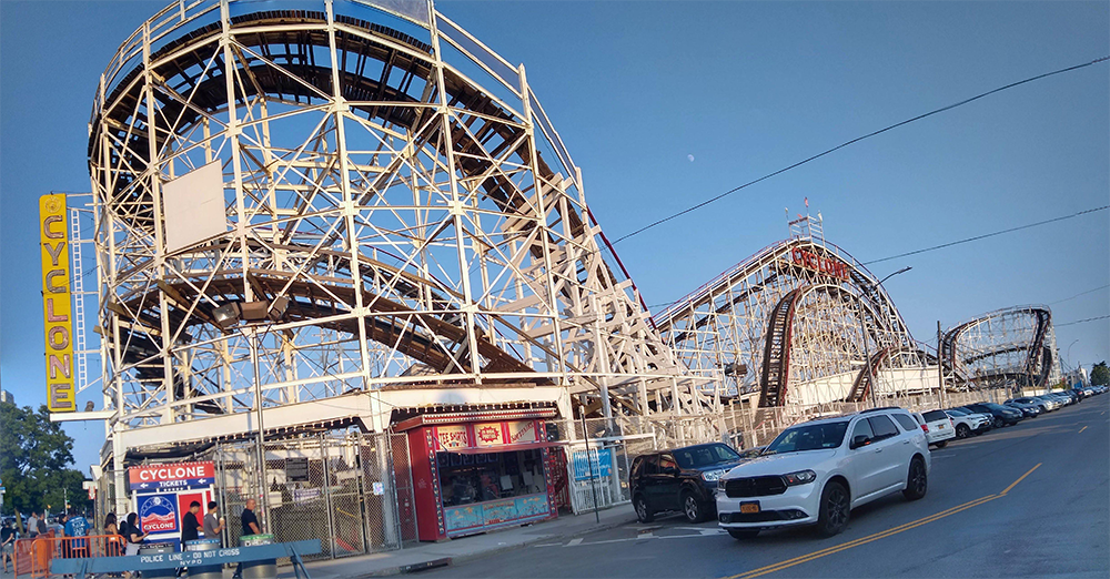 A large wooden roller coaster called Cyclone with white supports and brown tracks sits next to a street with parked cars under a clear blue sky. A ticket booth and Cyclone sign are visible in the foreground.