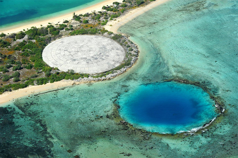 Aerial view of a tropical island with a large concrete dome structure on the shore and a circular deep blue water hole nearby in the turquoise shallow sea.