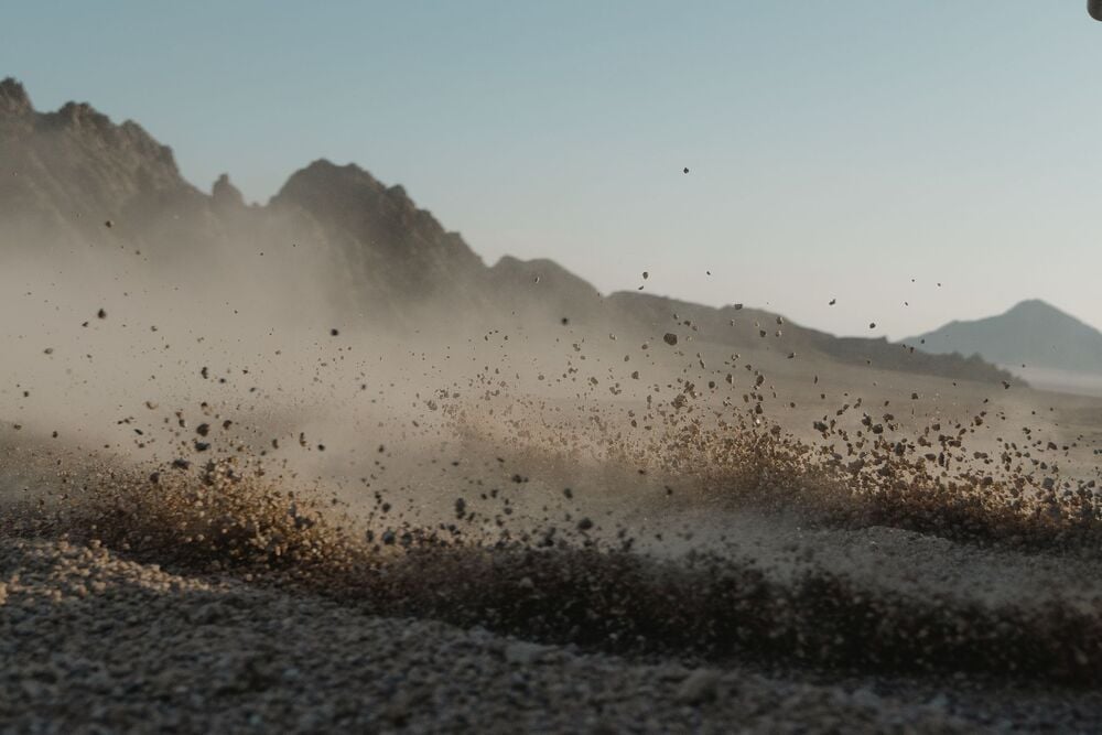 Dust and small rocks are kicked up from a dry, rocky ground with mountains in the background under a clear sky.