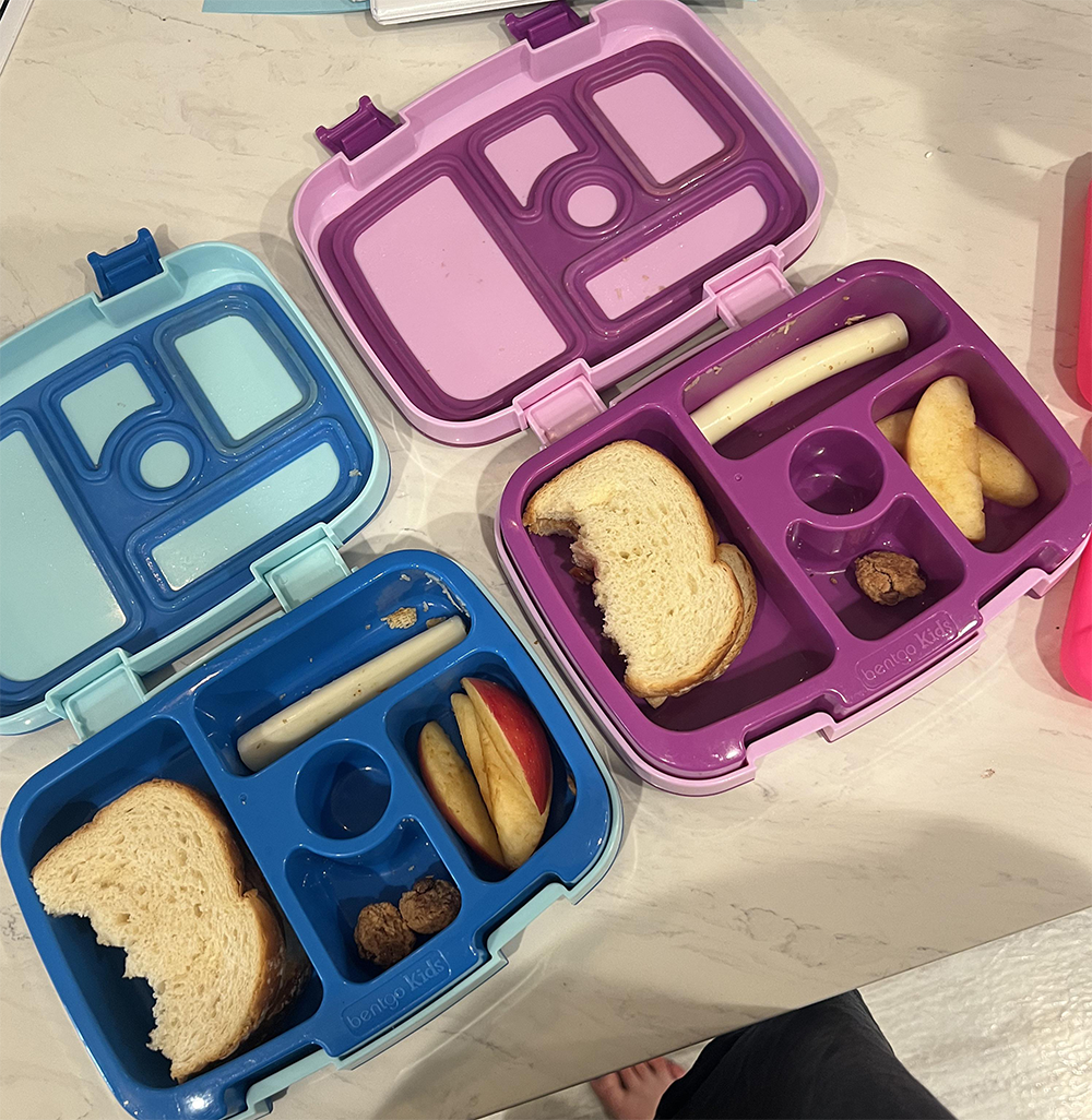 Three open lunchboxes with sandwiches, cheese sticks, sliced apples, and meatballs, placed on a light countertop. Each box is partially filled, two are blue and one is purple. A person’s feet are visible at the bottom.