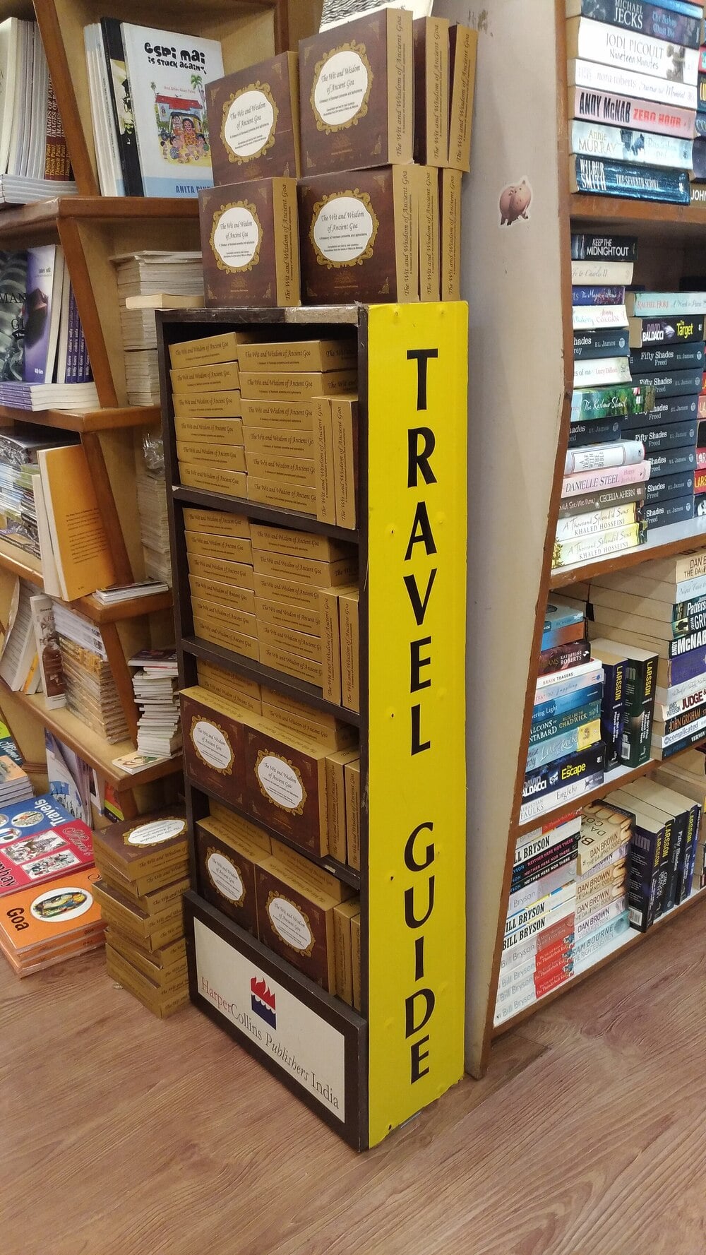 A bookstore display with a yellow “TRAVEL GUIDE” sign holds stacks of brown travel books, with more books and shelves seen in the background. The floor is wooden.