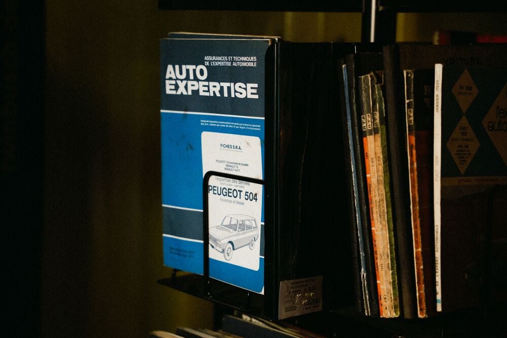 A blue and white “Auto Expertise” manual for a Peugeot 504 stands upright on a shelf among other books and magazines, with part of a car illustration visible on the cover.