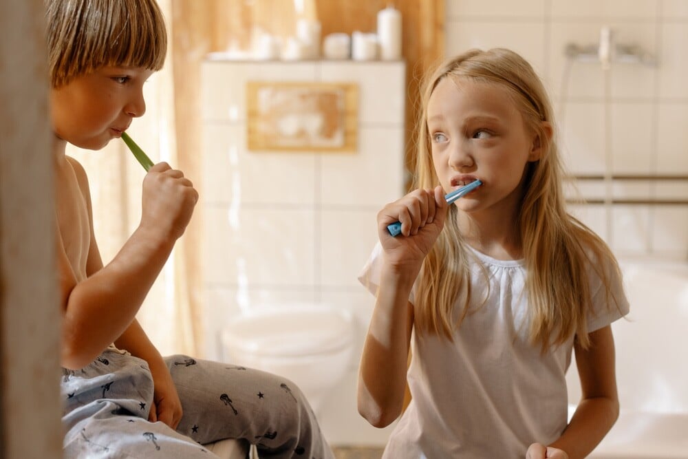 Two children in pajamas are brushing their teeth together in a bathroom with white tiles and soft natural light.