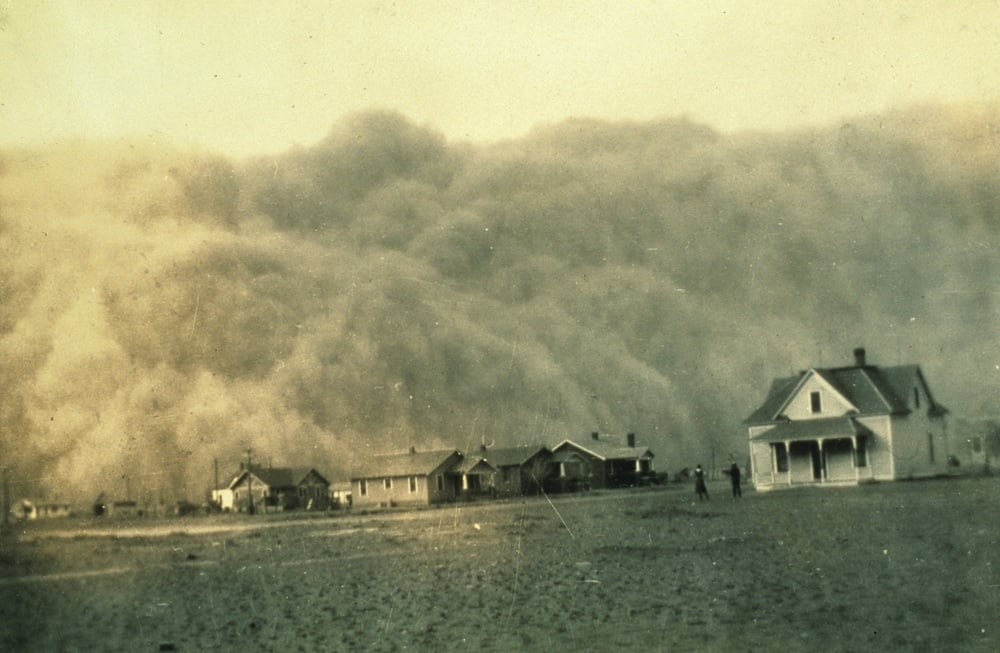 A massive dust storm approaches a row of houses across a barren, dry landscape, with a few small figures standing outside.