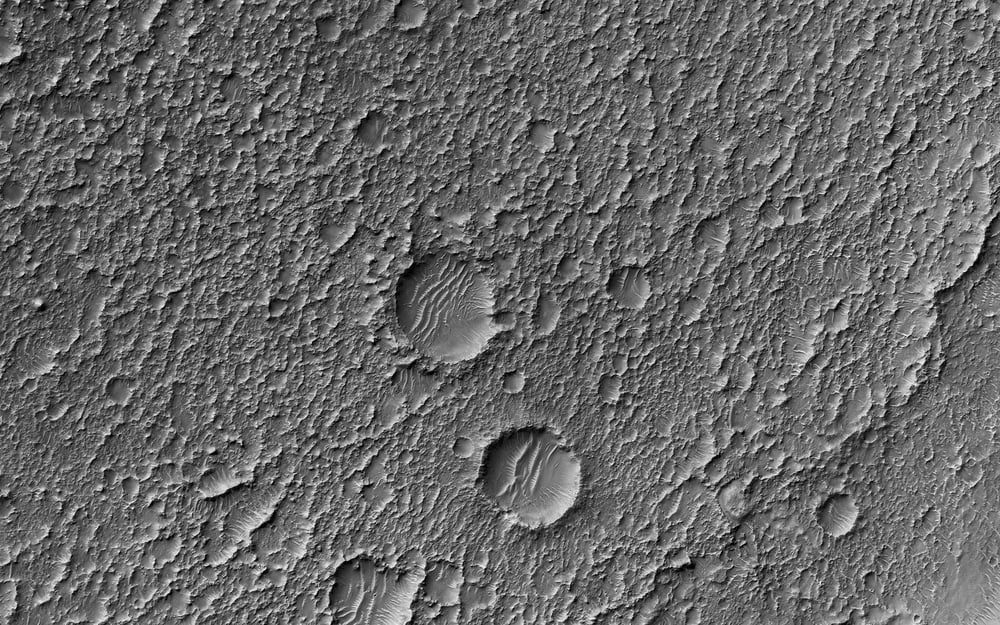A grayscale aerial view of a rocky, cratered surface with several round impact craters of varying sizes scattered across a rough, textured terrain.