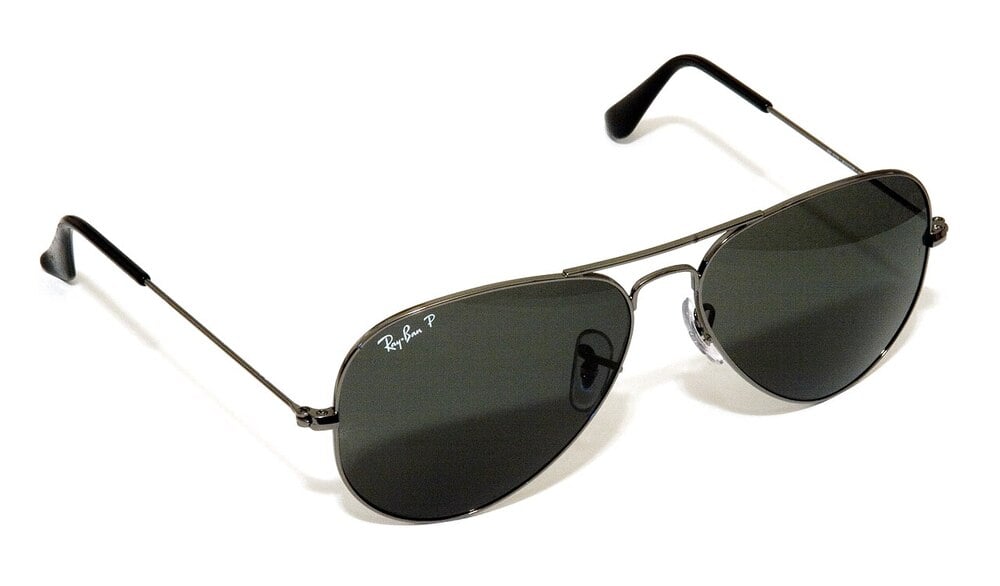 Black aviator sunglasses with dark lenses, thin metal frame, and black earpieces. The “Ray-Ban” logo is visible on the top left lens. The glasses are set against a white background.