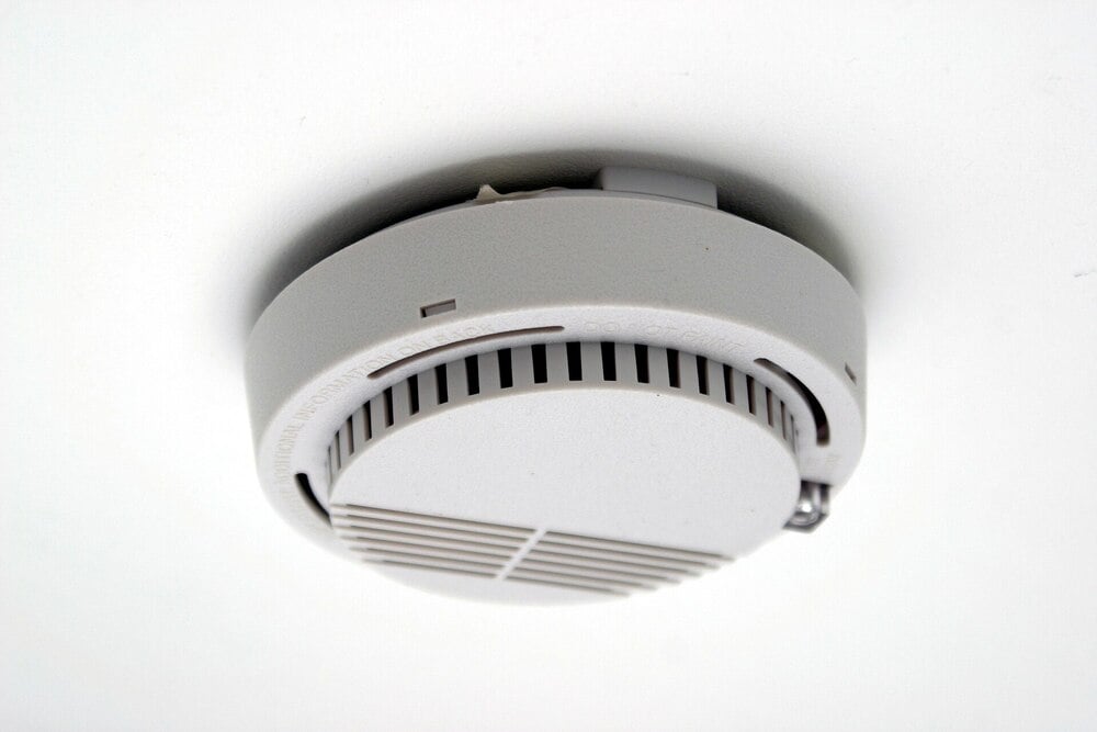 A white smoke detector mounted on a white ceiling, viewed from below. The device has ventilation slats and a small indicator light.