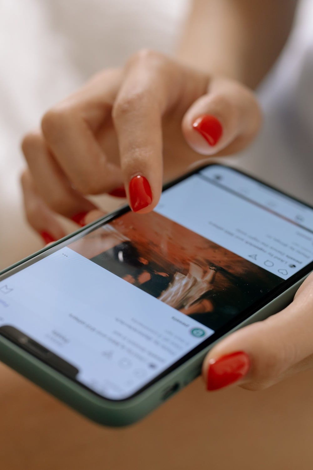 A person with red nail polish uses a smartphone, scrolling through a social media feed that displays an image featuring warm, brownish tones.