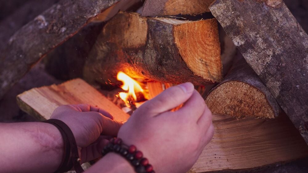 A person’s hands shield a small campfire made of stacked logs and kindling, helping the flame catch. The hands wear a brown bracelet and red bead bracelet.