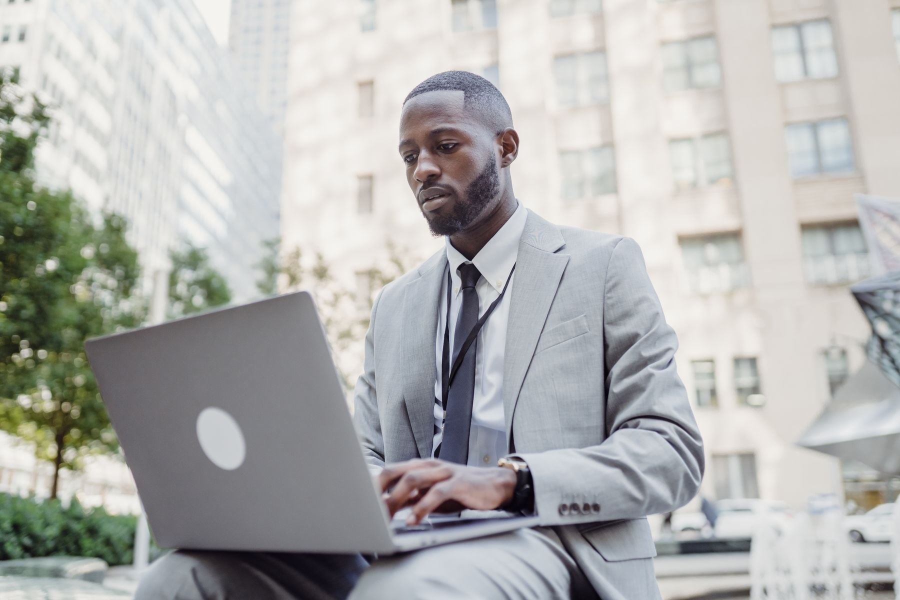 A man in a gray suit works on a laptop outdoors in an urban setting, with tall buildings and greenery in the background.