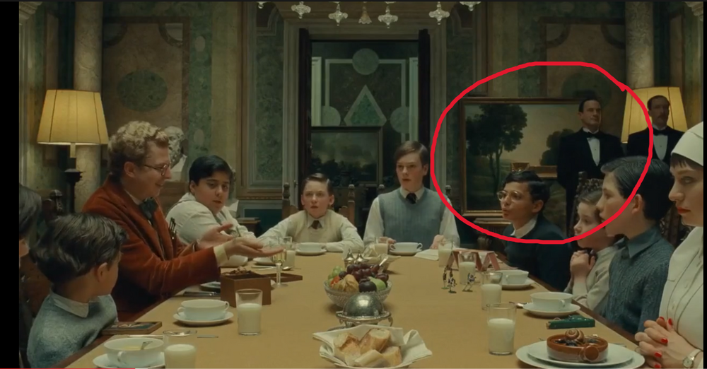 A group of people sits around a dining table set for a meal; two men circled in red stand and sit near a painting in the background.