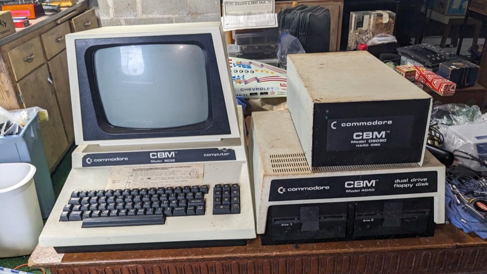 A vintage Commodore computer setup with a monitor, keyboard, dual floppy disk drives, and additional hardware, all labeled with CBM branding, sits on a wooden table in a cluttered room.