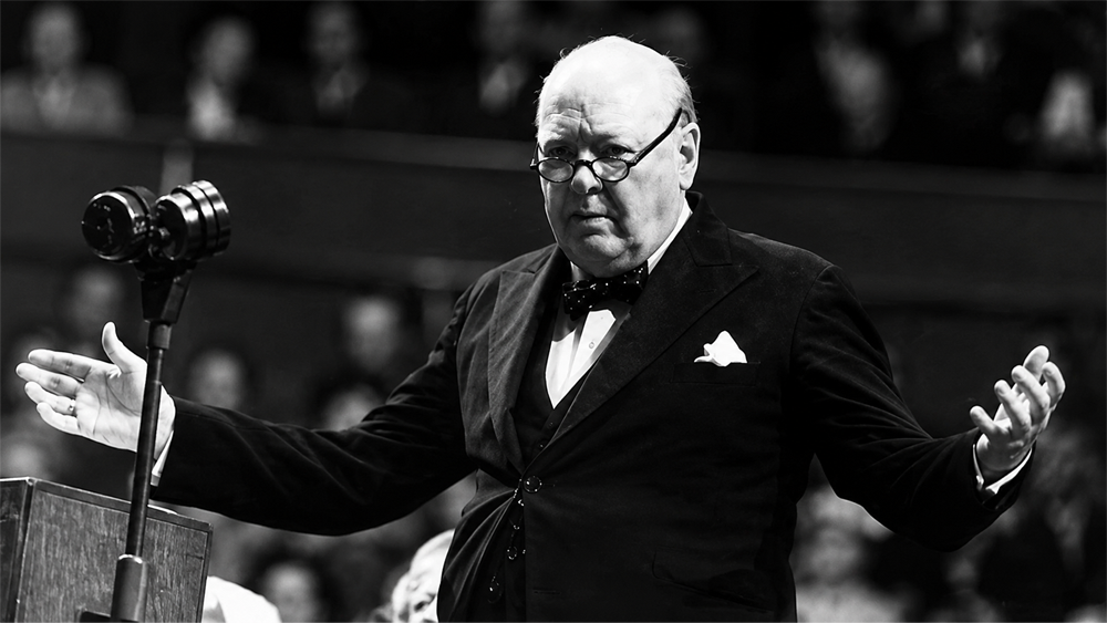 A man in a suit and bow tie stands at a podium with a microphone, arms outstretched, appearing to address an audience.