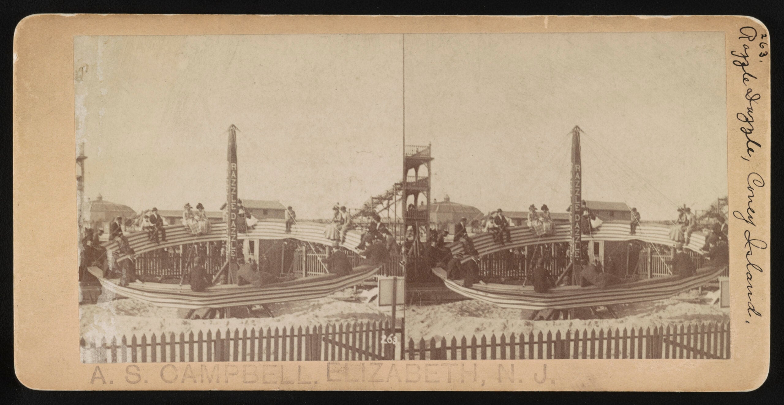 Vintage photo of people sitting on large wooden boats at an amusement park ride, with a picket fence in front and buildings in the background. The image is sepia-toned and labeled “A. S. CAMPBELL, ELIZABETH, N.J.”