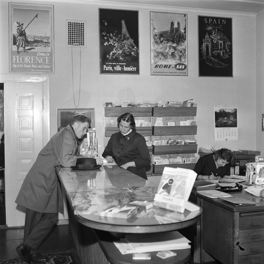 A man leans on a counter, speaking with a woman behind it, while another woman writes at a desk. Travel posters and brochures are visible in the background, suggesting a travel agency office.