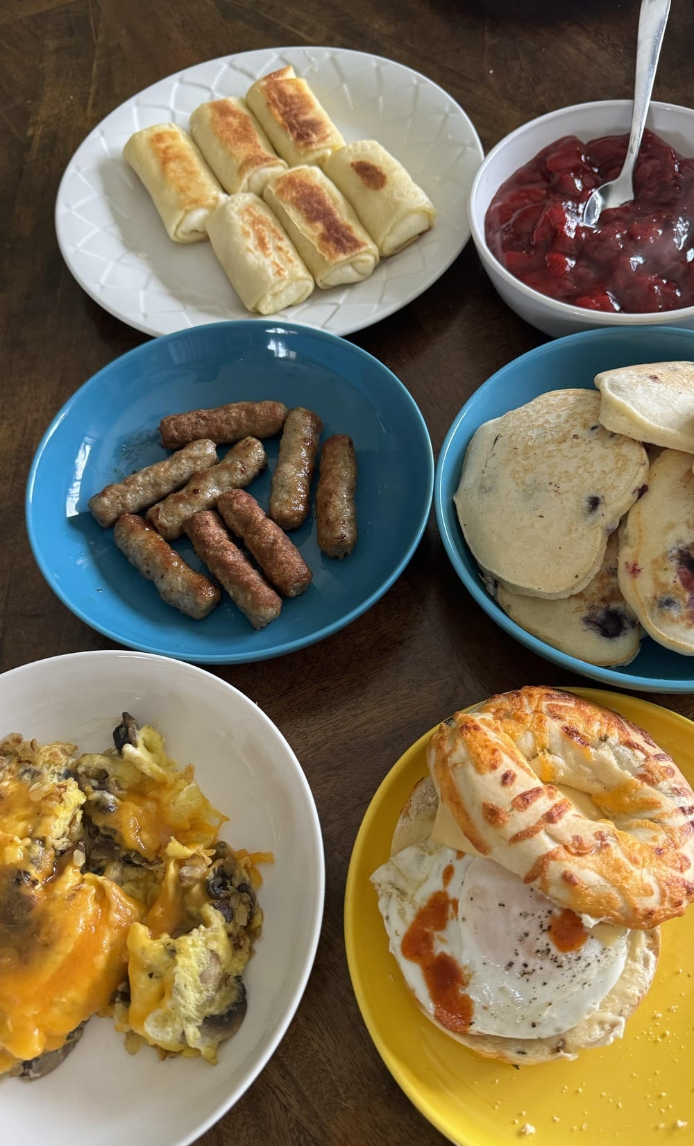 A table set with plates of blintzes, cherry topping, sausage links, blueberry pancakes, cheesy scrambled eggs, and a bagel sandwich with egg and hot sauce.