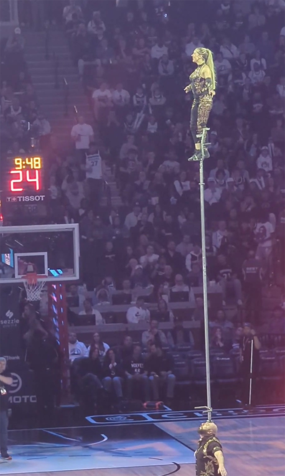 A performer balances high on top of a tall pole at a basketball game, with the audience watching and the shot clock showing 24 seconds and 9:48 on the scoreboard in the background.