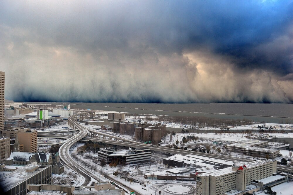 A snowy cityscape with roads and buildings is shown beneath dramatic, dark storm clouds approaching over a large, partially frozen body of water.
