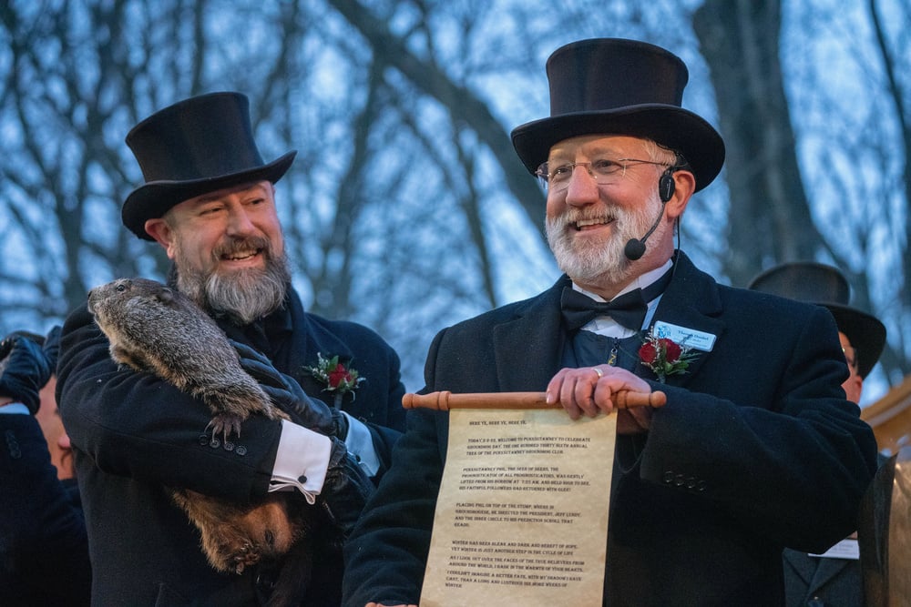Two men in black top hats and coats stand outdoors. One holds a groundhog; the other holds a scroll and wears a headset microphone. Both are smiling, and bare trees are visible in the background.