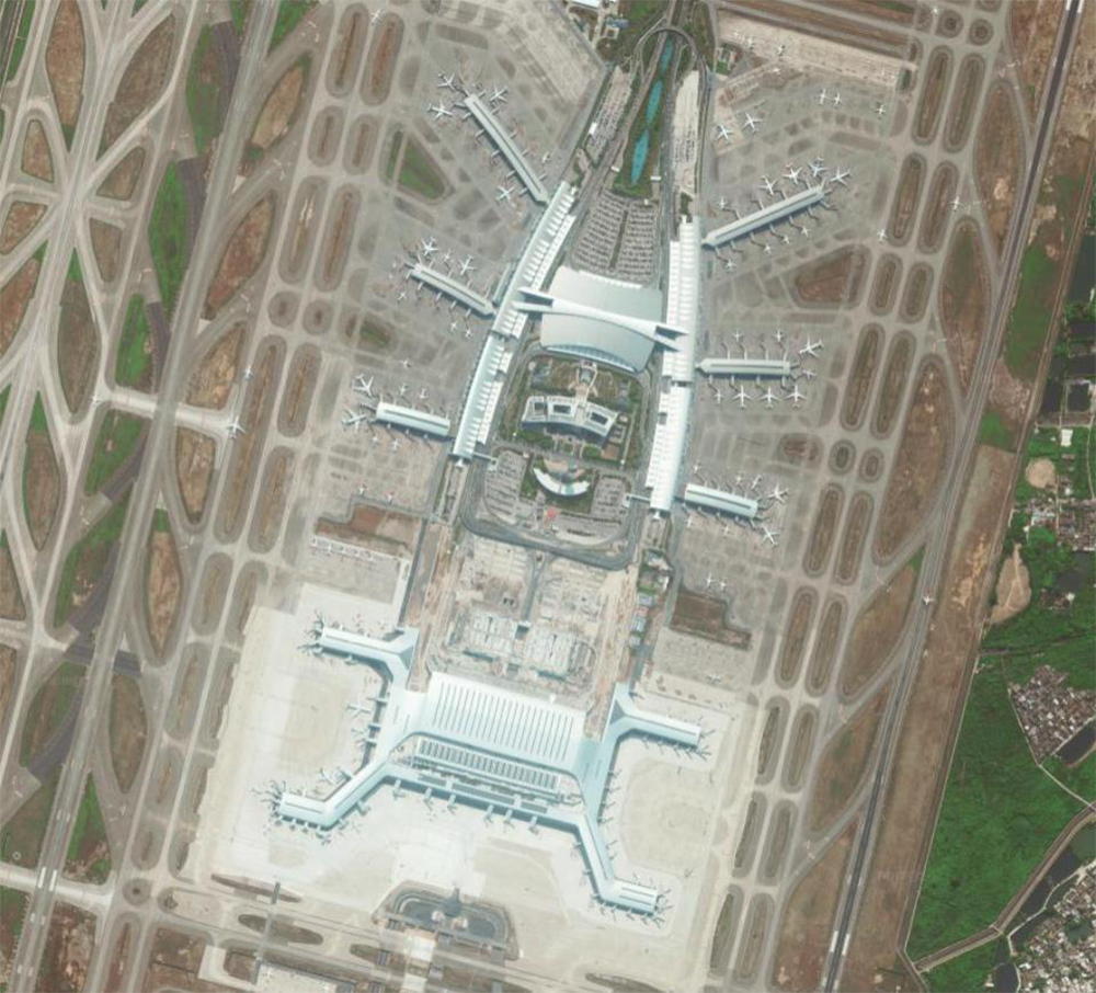 Aerial view of a large, modern airport terminal with multiple gates, airplanes parked around it, and several runways and taxiways visible. Nearby, there are patches of greenery and a small urban area.