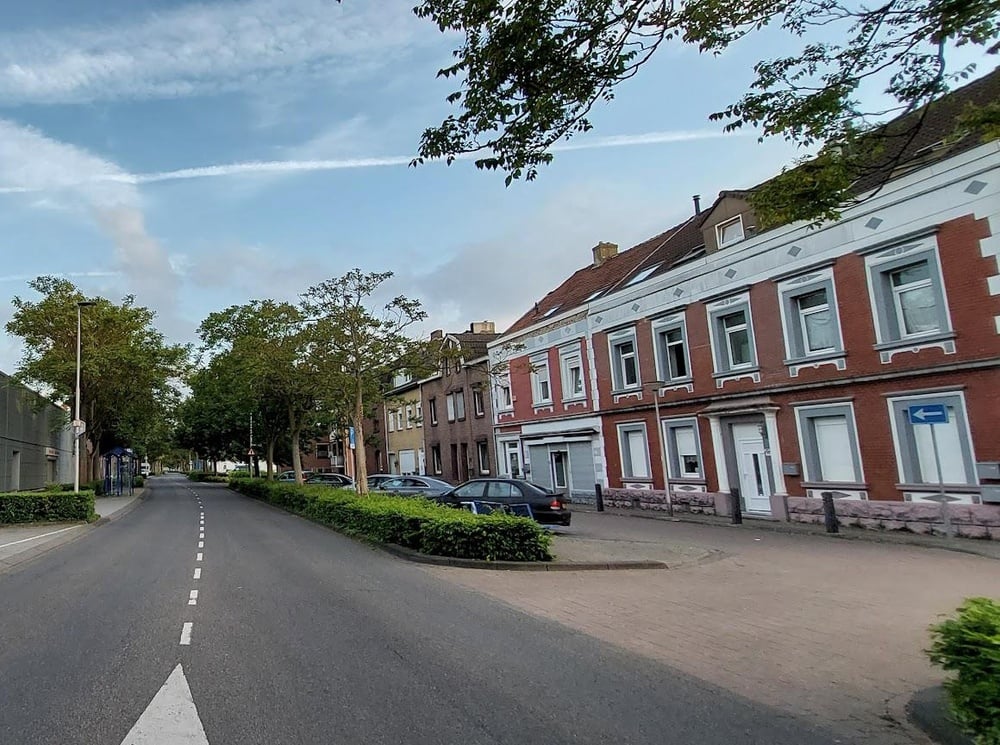 A quiet street with trees and parked cars beside old-style brick buildings under a partly cloudy sky. A blue one-way traffic sign is visible near the buildings.