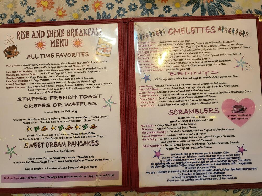A laminated breakfast menu with sections for all-time favorites, pancakes, omelettes, and scramblers, featuring colorful headings and various breakfast dish descriptions on a floral tablecloth.