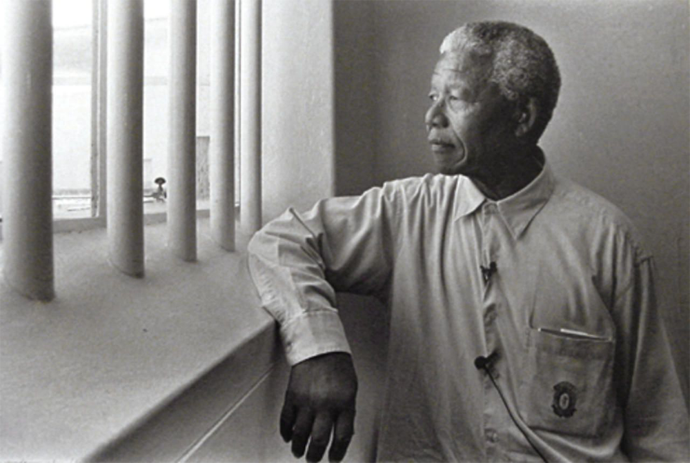 An older man with gray hair stands by a barred window, looking outside thoughtfully. He wears a buttoned shirt and has a calm, reflective expression.