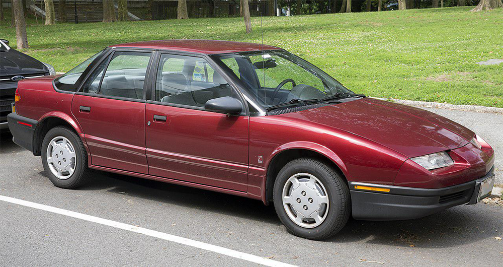 A red four-door sedan is parked by a curb in a residential area with grass and trees in the background. The car has silver hubcaps and black trim.