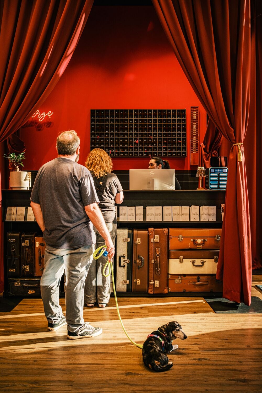 A man with a small dog on a leash stands in a hotel lobby with red curtains, while a woman speaks to the receptionist at the front desk made of stacked vintage suitcases.