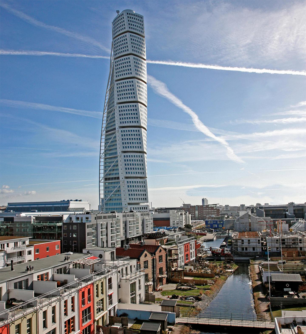 A tall, twisting skyscraper rises above a cityscape with colorful buildings, a canal, and blue sky with white contrails.