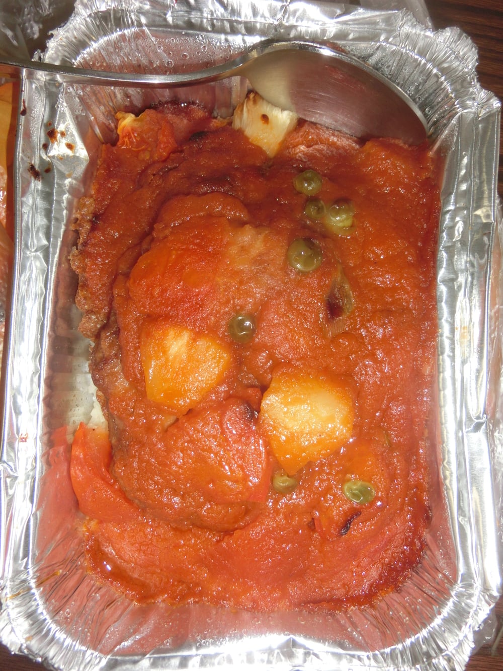 A foil container of food with tomato sauce, potatoes, peas, and a spoon in one corner.