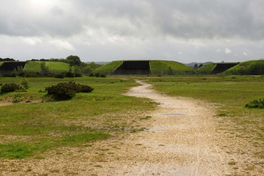 A gravel path leads across a grassy field toward two grass-covered bunkers under a cloudy sky. Shrubs and trees are scattered in the background.