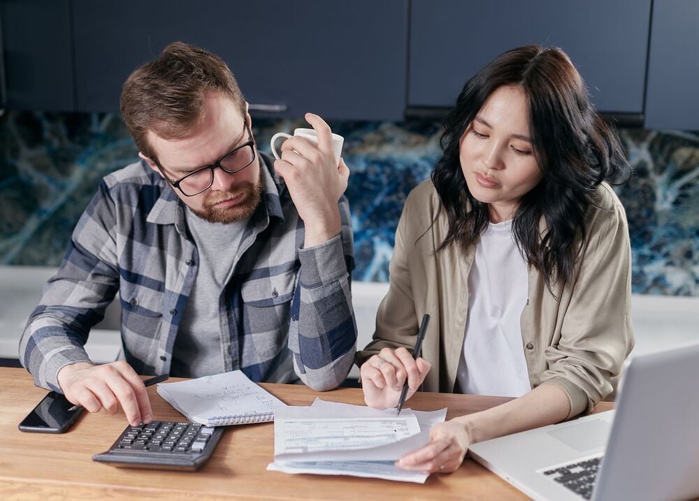 A man and woman sit at a table with papers, a calculator, and a laptop, appearing focused as they review finances or bills together.