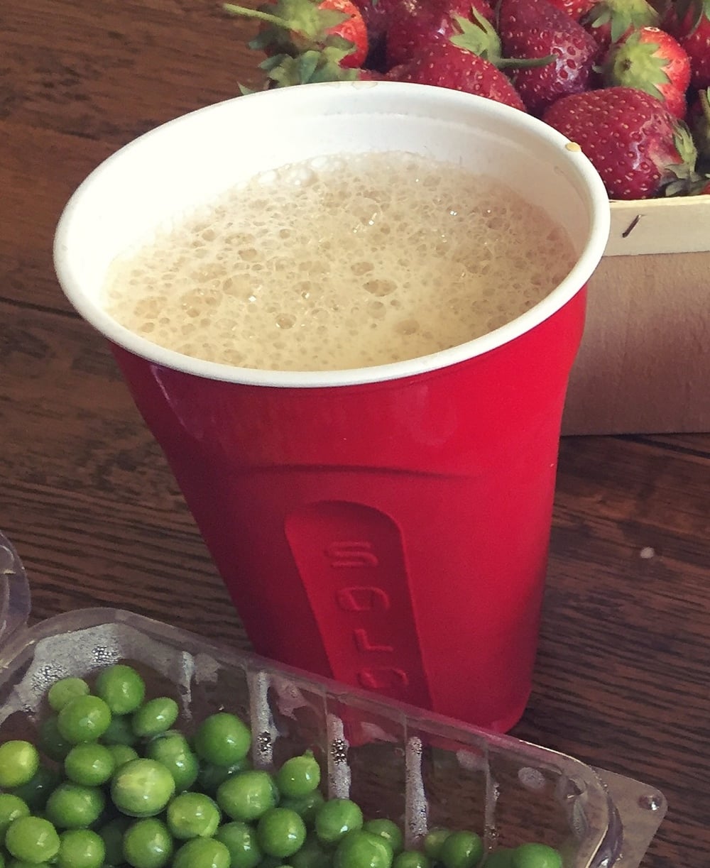 A red plastic cup filled with a frothy drink sits on a wooden table near a basket of strawberries and a container of green peas.