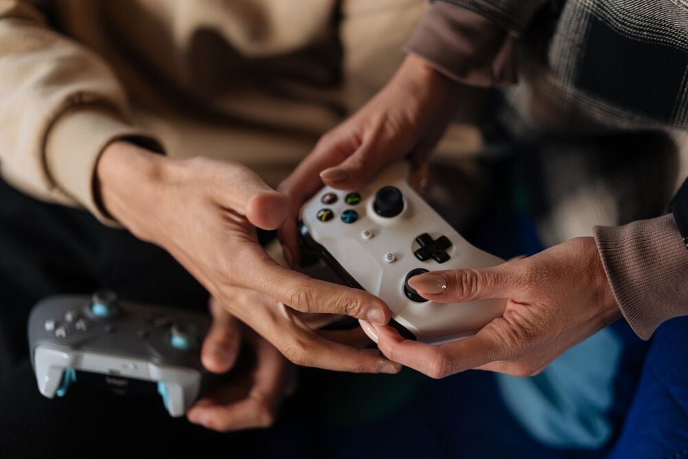 Two people sit close together, holding and playing with video game controllers. One person holds a white controller while the other holds a gray one, both focused on the game.