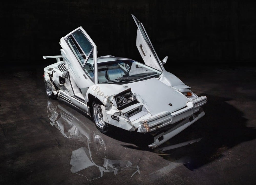 A damaged white Lamborghini sports car with its scissor doors open, sitting on a reflective surface in a dark setting. The front and side panels are visibly dented and scratched.
