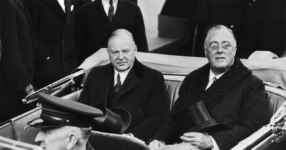 Two men in suits sit in the back of an open car during a formal event, with one holding a top hat. Other men in suits are standing around them.