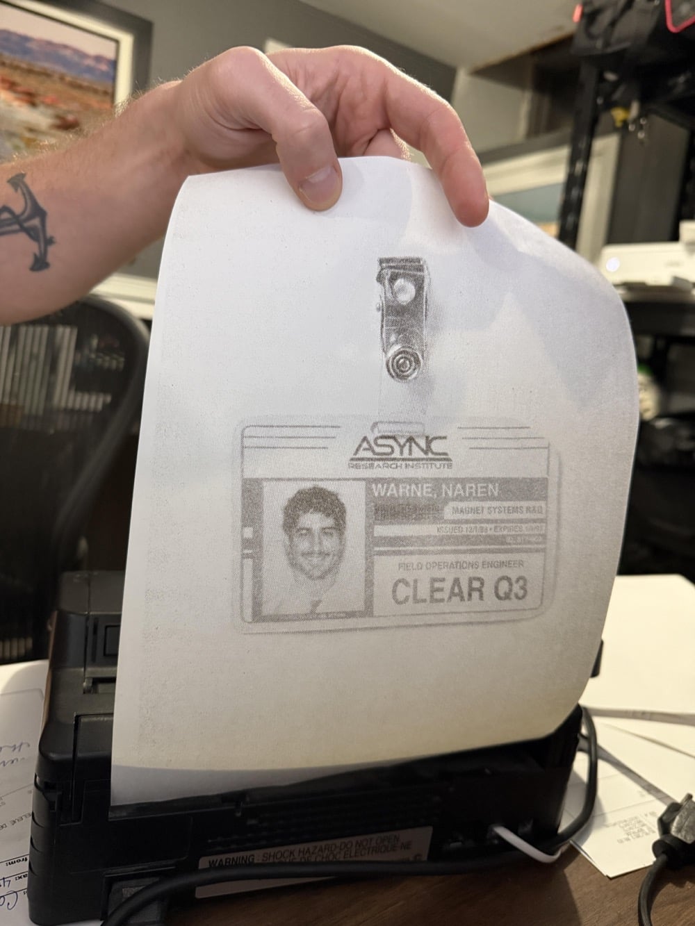 A hand pulls a paper from a small printer. The paper shows a grayscale copy of an ID badge with a photo, text, and the label “CLEAR Q3.” Office supplies are visible in the background.