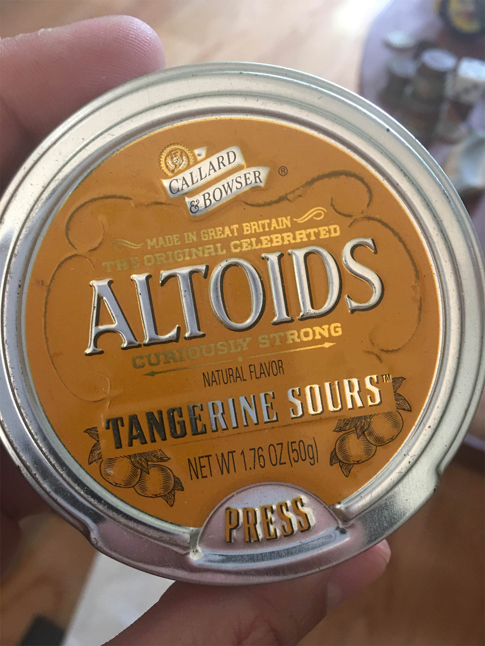 A hand holds a round tin of Callard & Bowser Altoids Curiously Strong Tangerine Sours, featuring an orange and silver design with a “Press” button at the bottom.