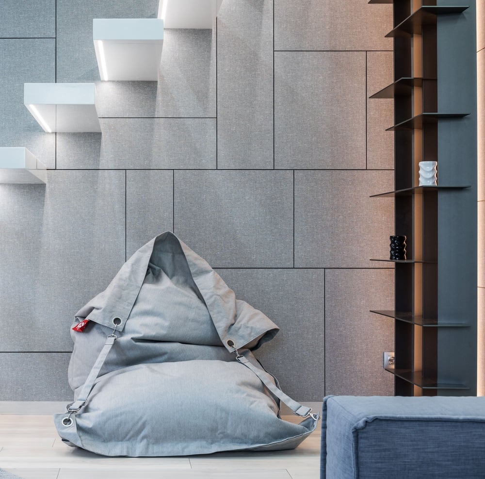 A large gray bean bag sits in front of a geometric wall with built-in shelves and stairs. The room has a modern, minimalist design with neutral tones.