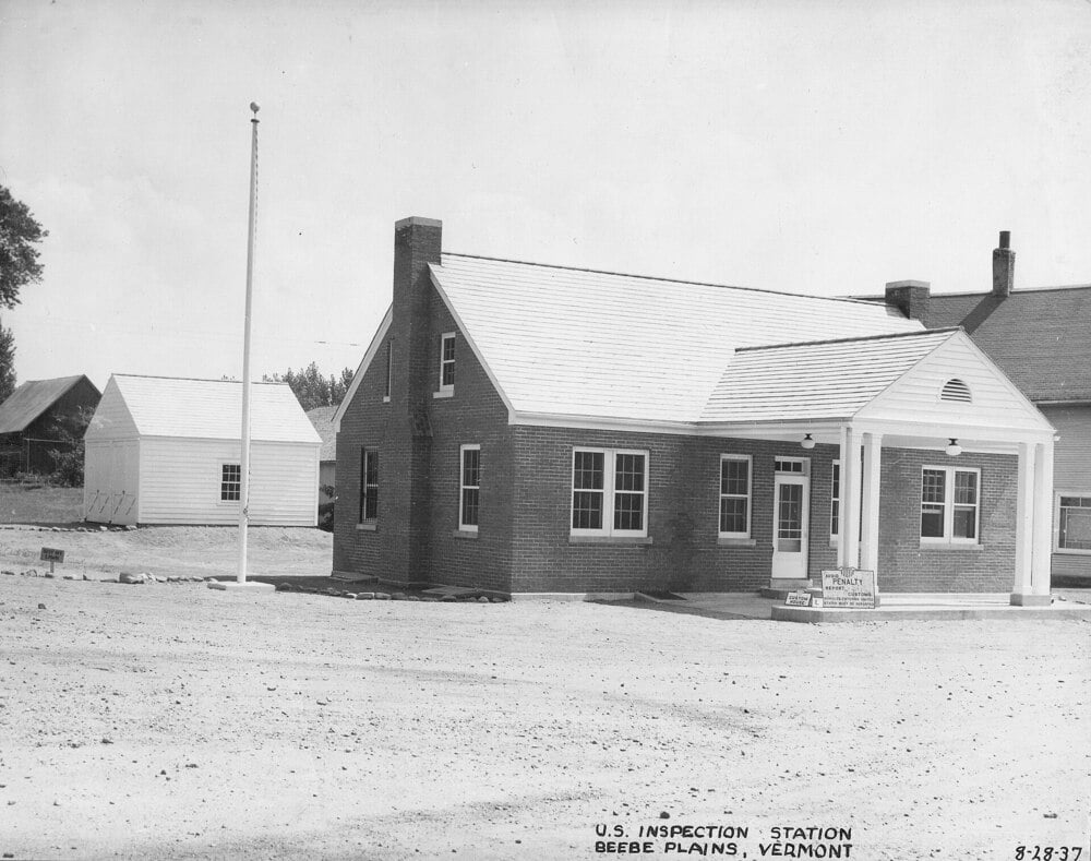 A brick building with a white porch labeled “U.S. Inspection Station” sits by a flagpole on a dirt lot in Beebe Plains, Vermont. Another small building stands in the background. The photo is dated 8-28-37.