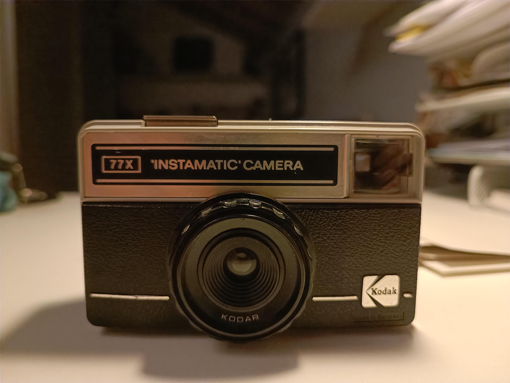 A close-up of a vintage Kodak Instamatic 77X camera sits on a white surface with blurred shelves and objects in the background.