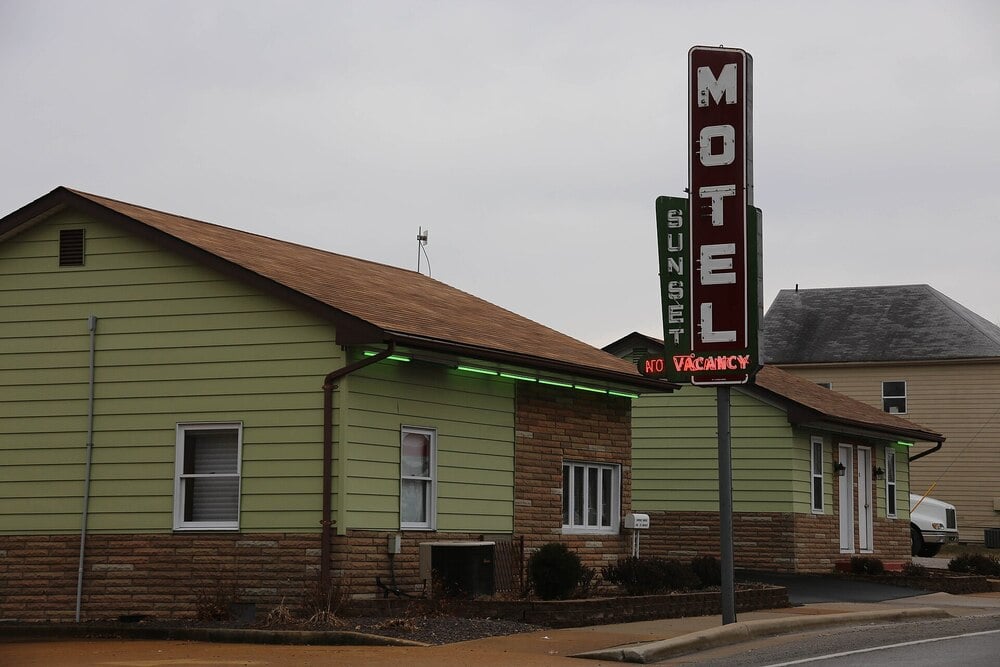 A small, single-story motel with pale green siding and a brown roof. A vertical sign reads “Sunset Motel.” The sign’s neon lights indicate “No Vacancy.” The weather appears overcast.